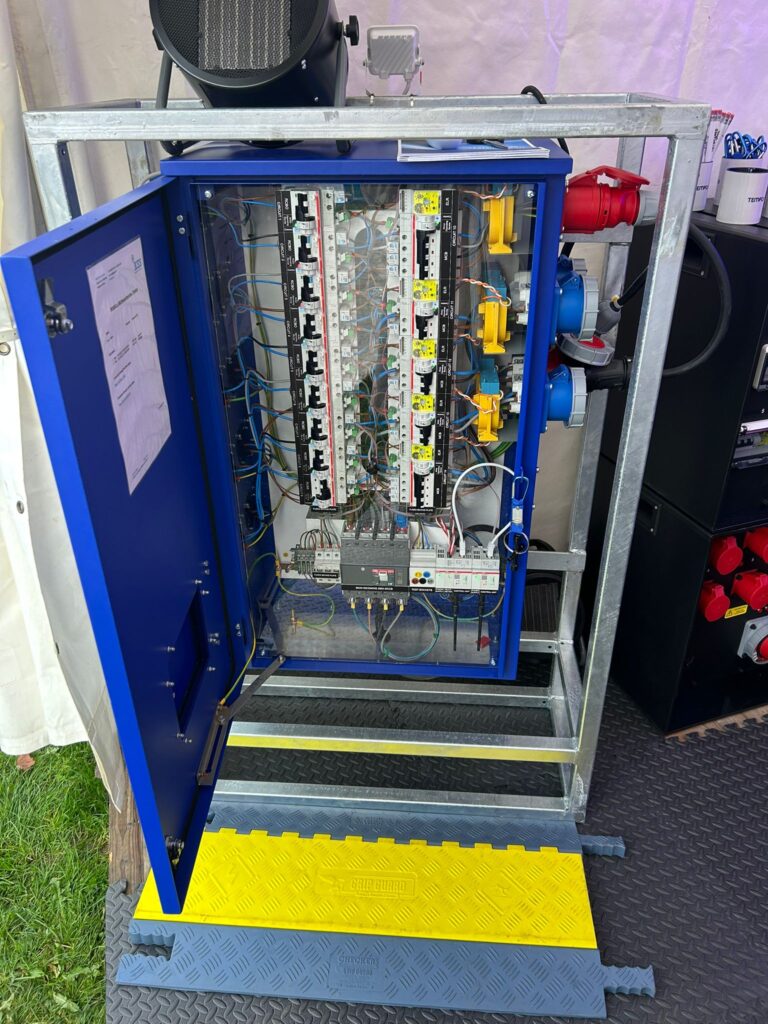 An open electrical control panel with multiple switches, wires, and connectors is mounted on a metal frame, with a yellow and blue cable protector on the ground in front.