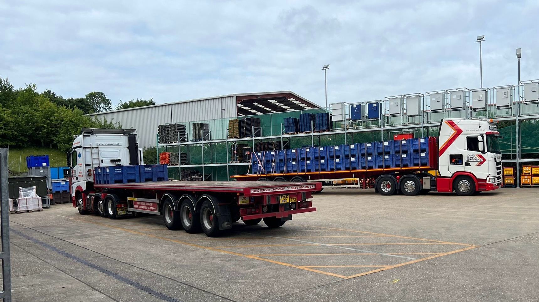 Two flatbed trucks are parked in a warehouse yard with stacked plastic containers and crates visible in the background.