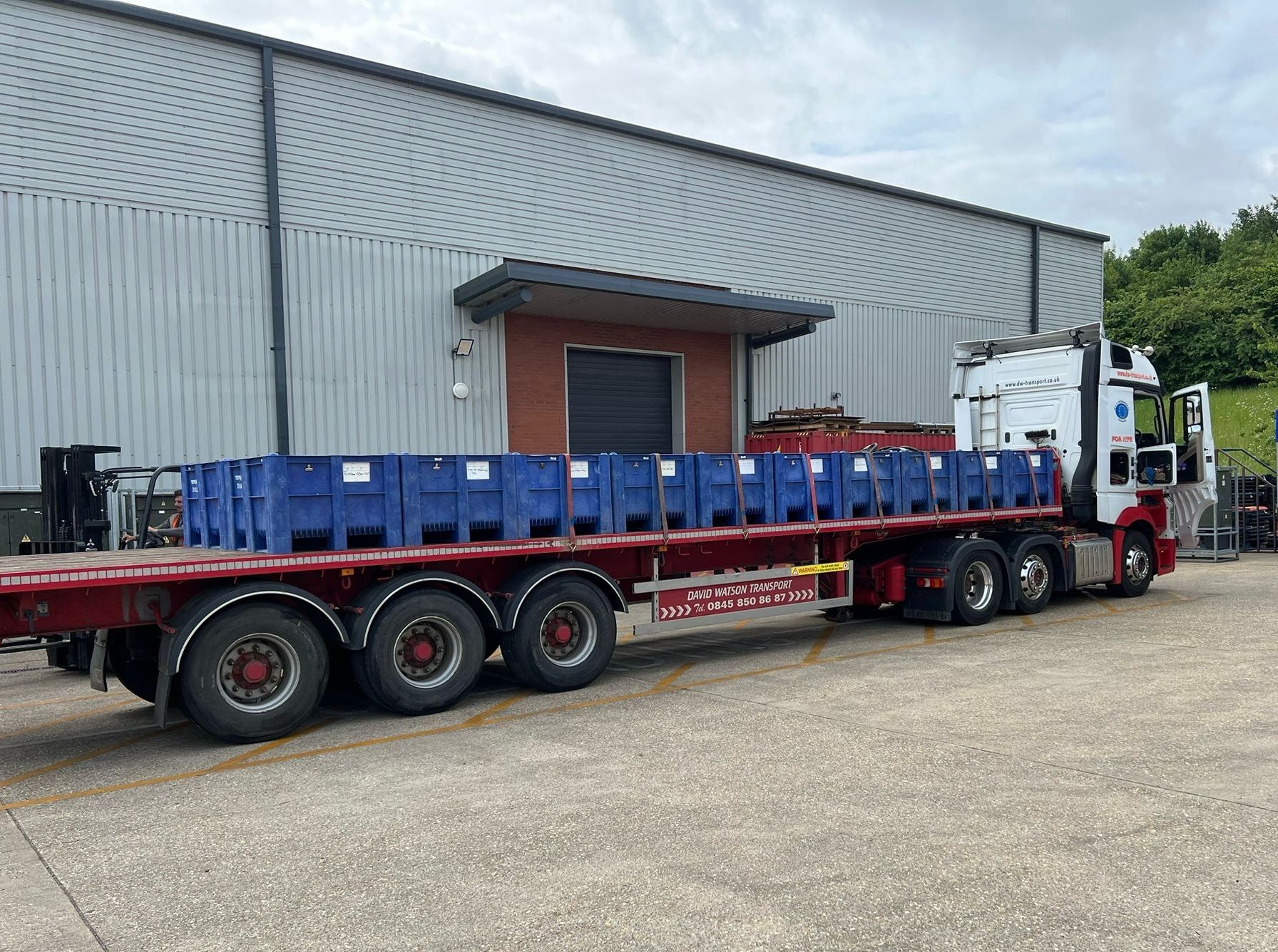 A flatbed truck parked outside a warehouse, carrying several large blue plastic containers secured on its trailer.