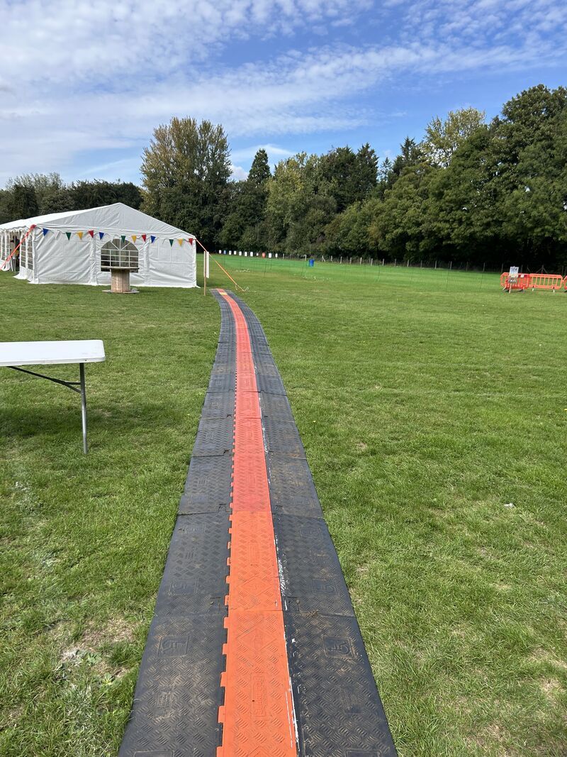 A temporary walkway with a red strip runs across a grassy field toward a white event tent, with trees and blue sky in the background.