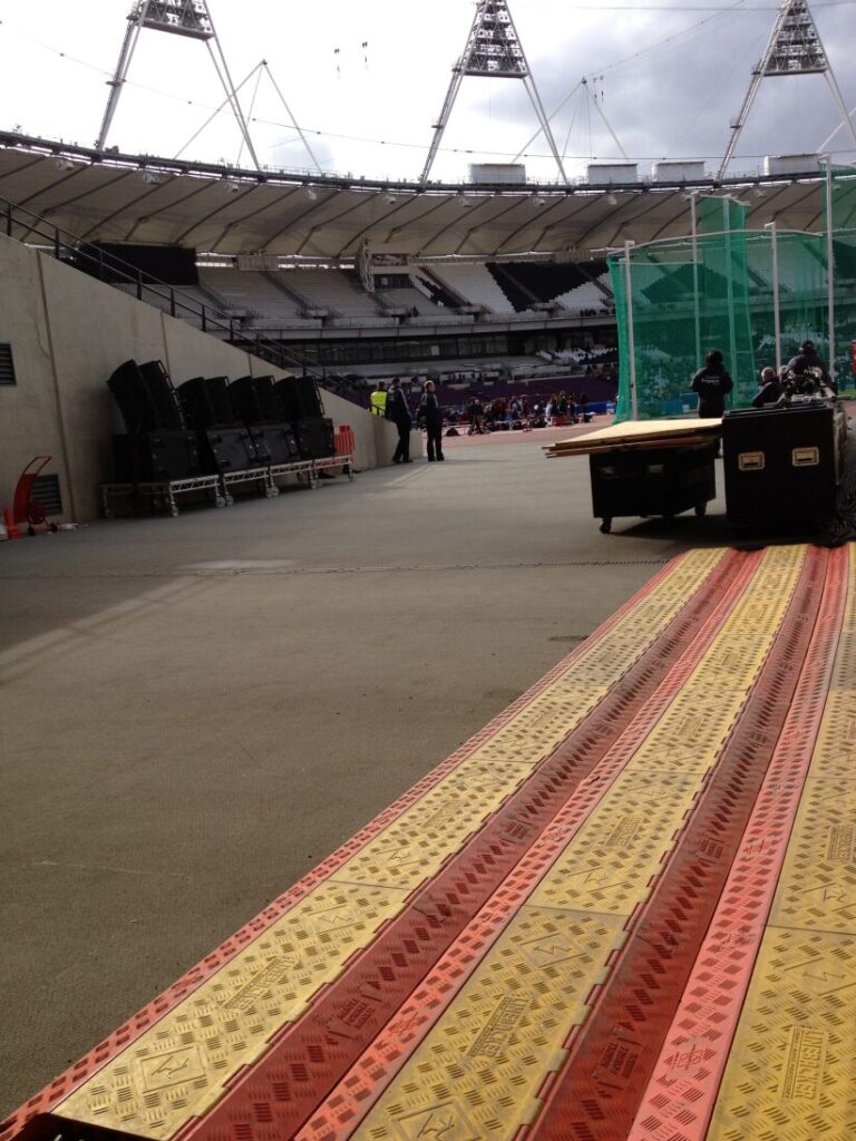 Several colored cable covers run along the ground inside a mostly empty stadium, with some people and equipment visible in the background.