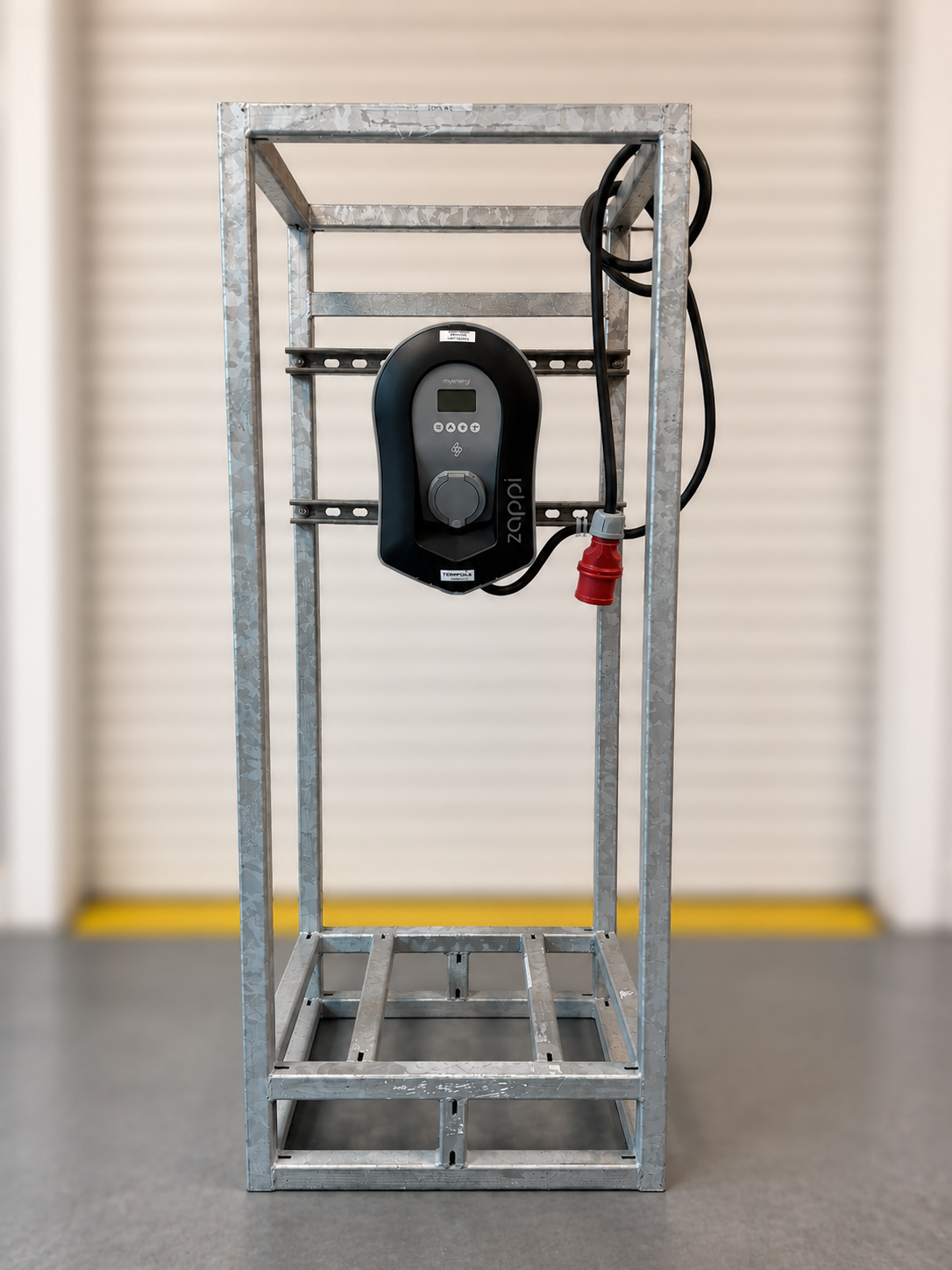 A black electric vehicle charging unit mounted in the center of a metal frame, with cables visible, set indoors on a gray floor.