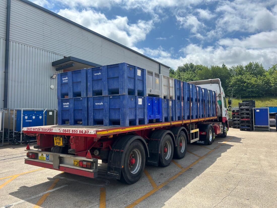 A flatbed truck loaded with blue and gray plastic crates is parked outside a warehouse on a sunny day.
