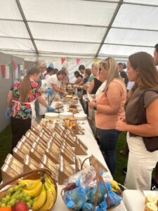 People gather inside a tent, lining up at a long table filled with baked goods, fruit, and packaged food items, with colorful bunting hanging in the background.