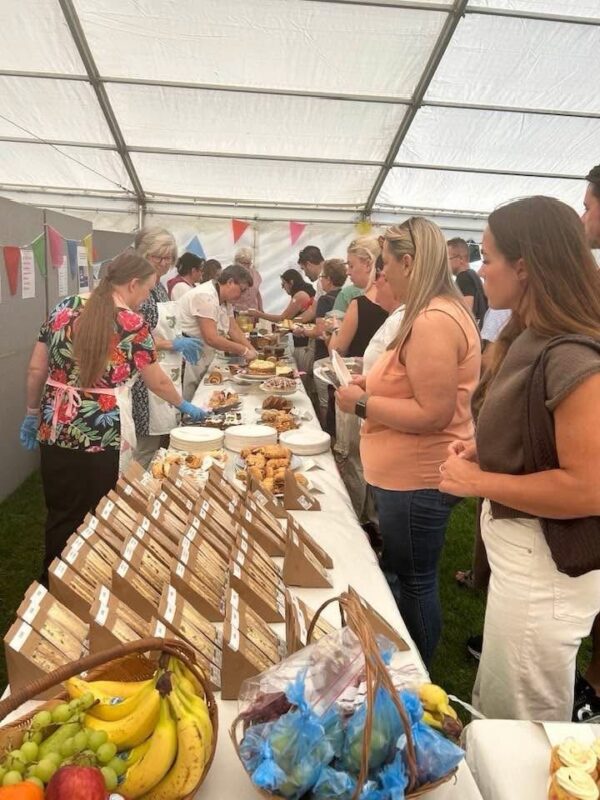 People gather inside a tent, lining up at a long table filled with baked goods, fruit, and packaged food items, with colorful bunting hanging in the background.